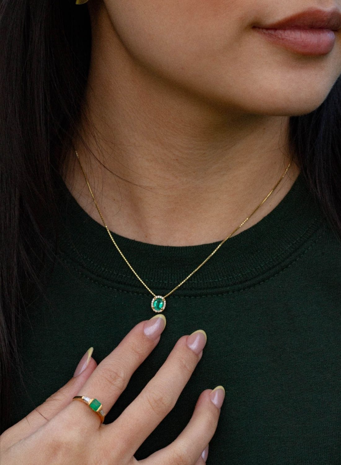Woman wearing a delicate gold necklace with a small oval emerald pendant, accented by diamonds and a simple, cushion cut emerald band ring.