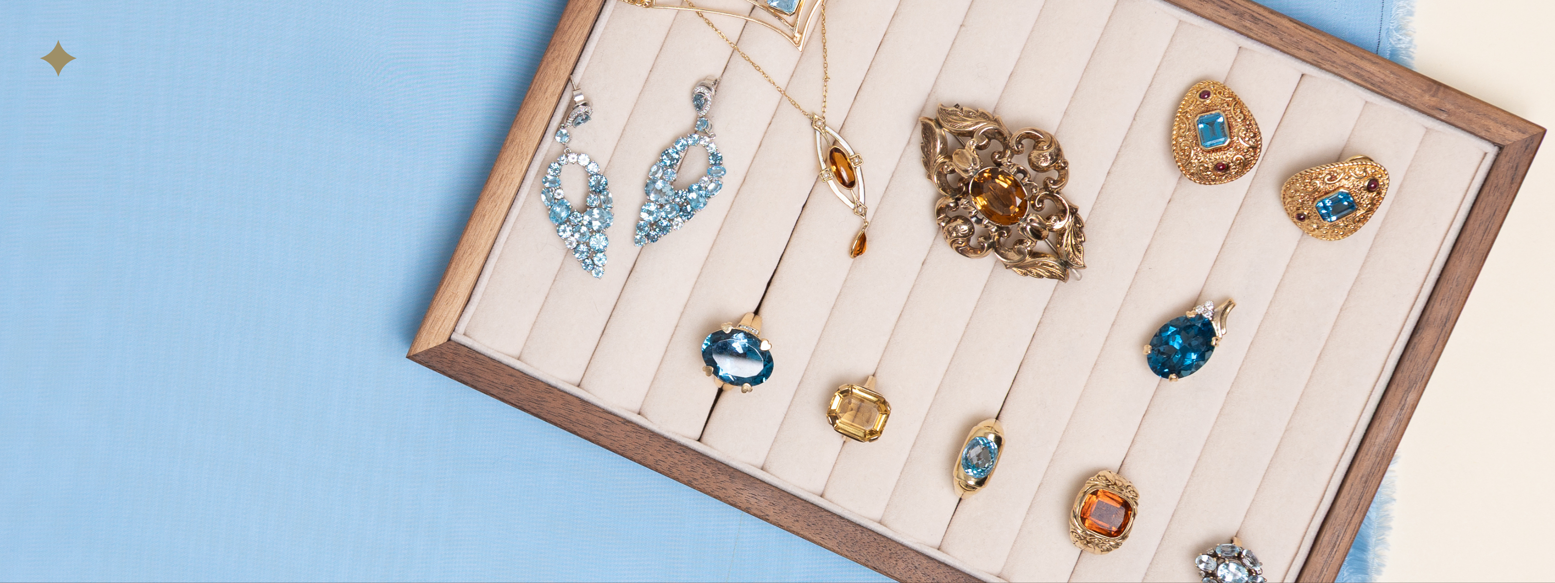 A collection of vintage citrine and blue topaz jewellery pieces arranged in a cream velvet tray with a wooden edge. The assortment includes gold rings, pendants, and brooches featuring oval, square, and teardrop-cut gemstones in warm yellow and cool blue tones, photographed on soft blue fabric.