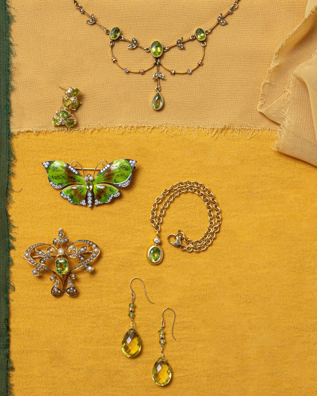 A jewelry set displayed on textured yellow chiffon, featuring a statement peridot delicate necklace, peridot drop earrings, a butterfly enamel brooch, a butterfly pin, and a gold chain with a oval peridot pendant.