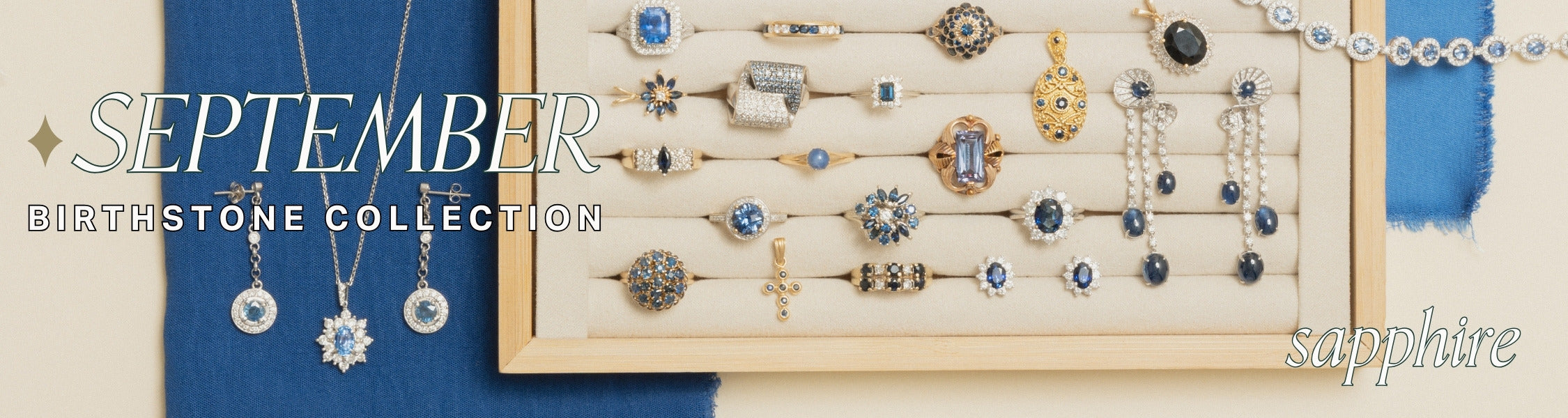Jewelry display on cream and wood jewellery tray over cobalt blue linen fabric. Assorted fine gold jewellery features a range of blue sapphire gemstone jewellery pieces with text overlay: September Birthstone Collection, sapphire