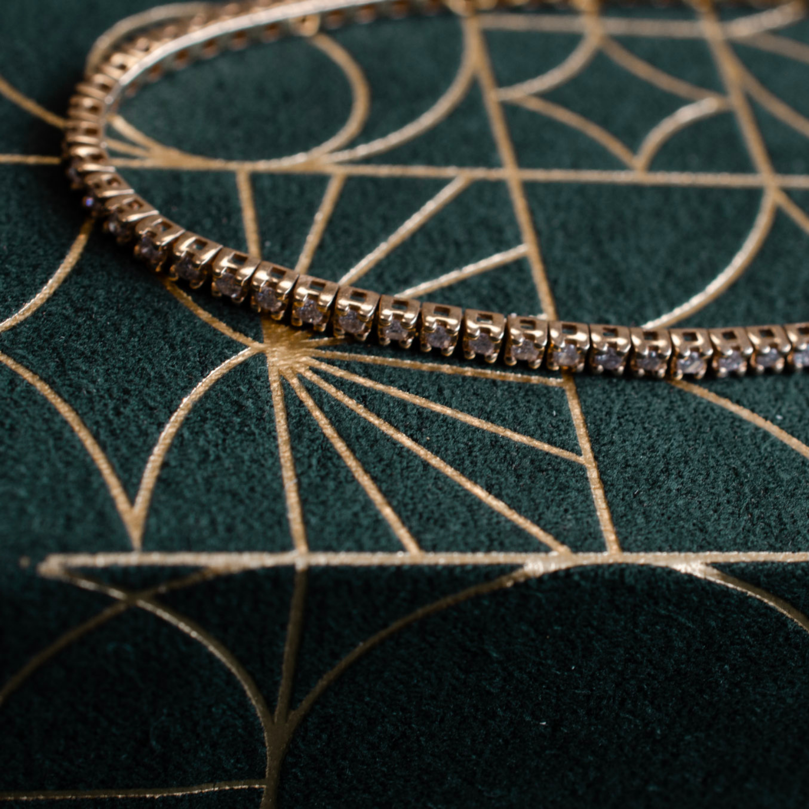Elegant secondhand gold diamond tennis bracelet resting on a rich dark green velvet fabric with a modern art deco gold pattern.