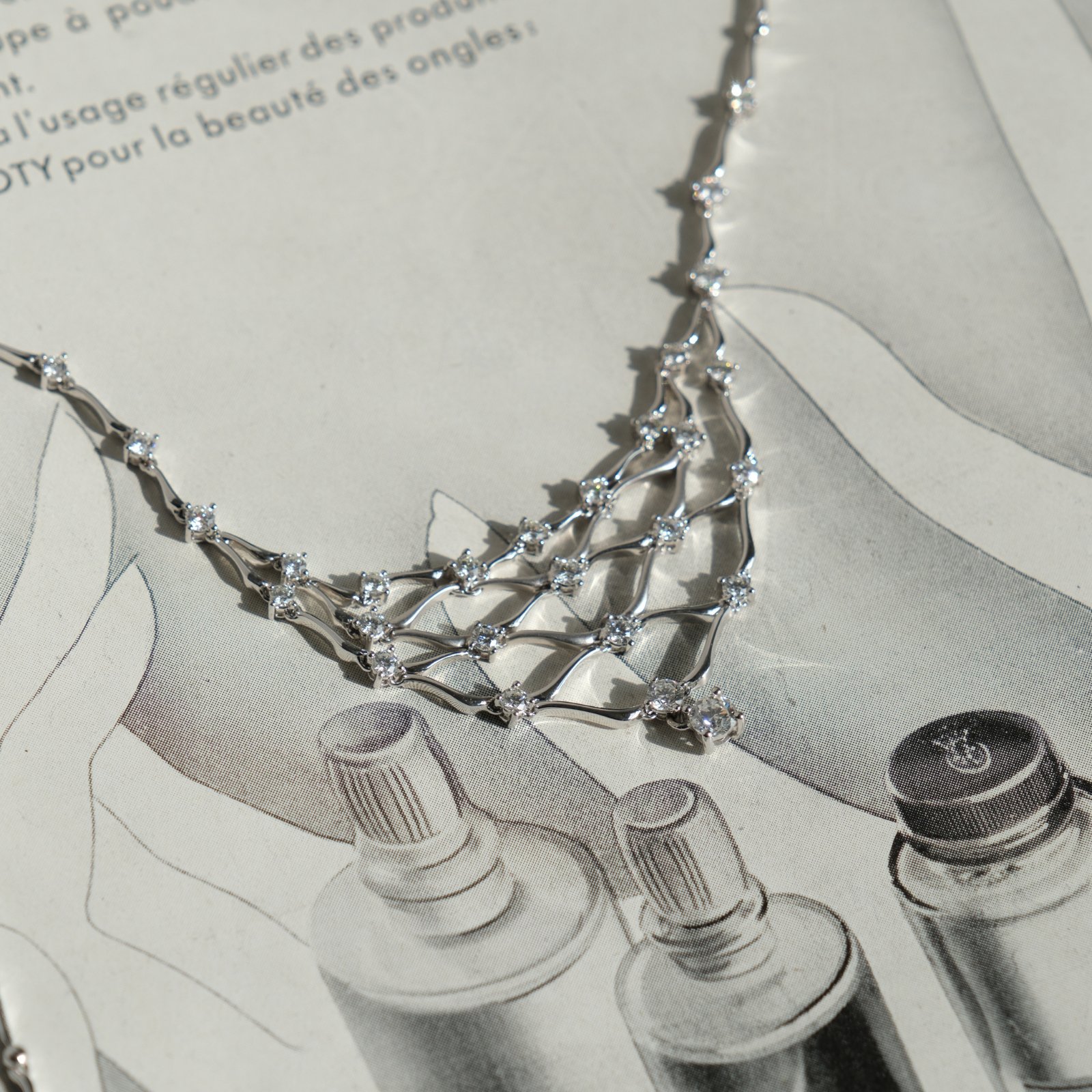 Delicate estate white gold and diamond draping bib necklace laid flat across a vintage black and white French fashion magazine page.