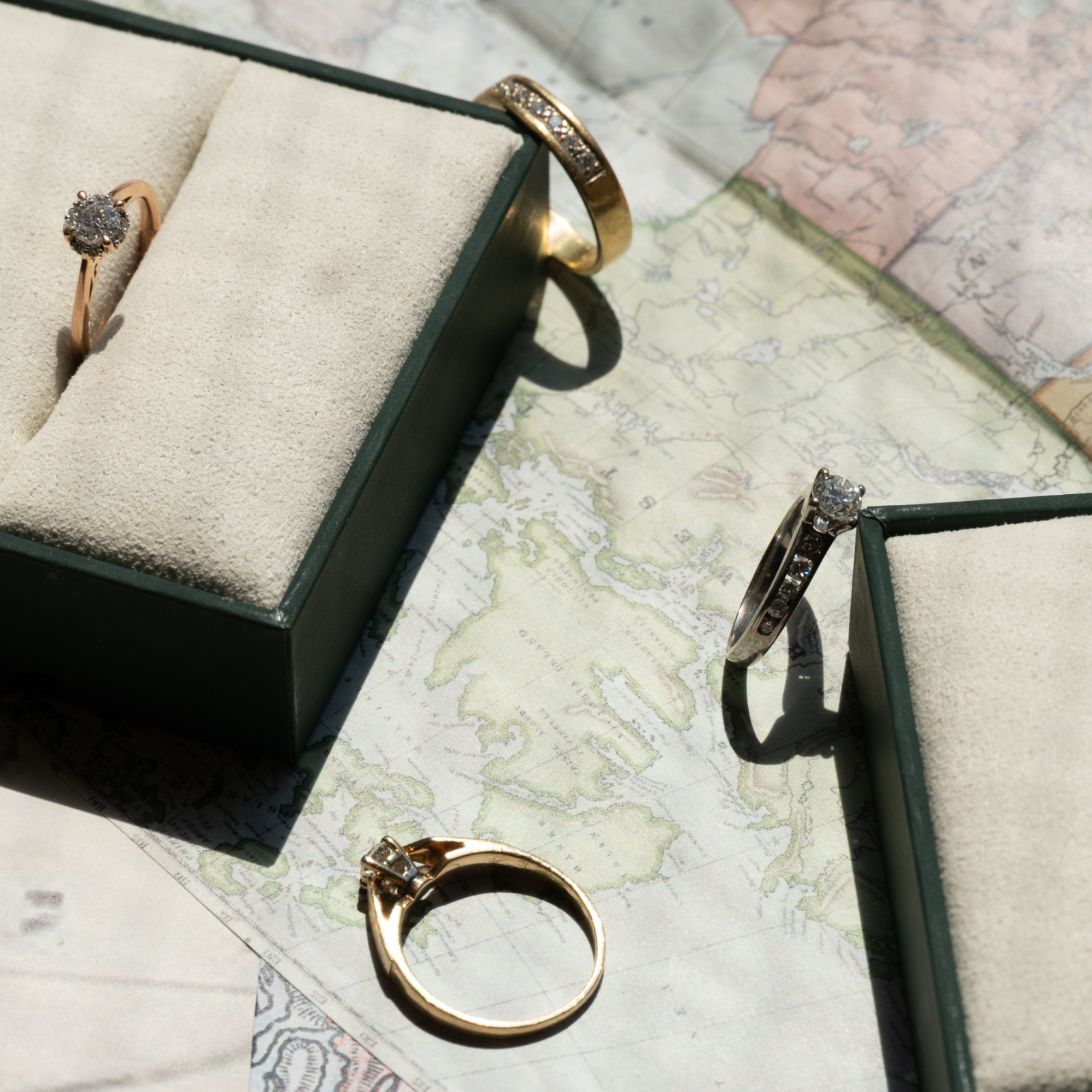 Curated collection of secondhand gold and diamond rings and jewellery bands displayed on an antique map alongside dark green velvet ring boxes.