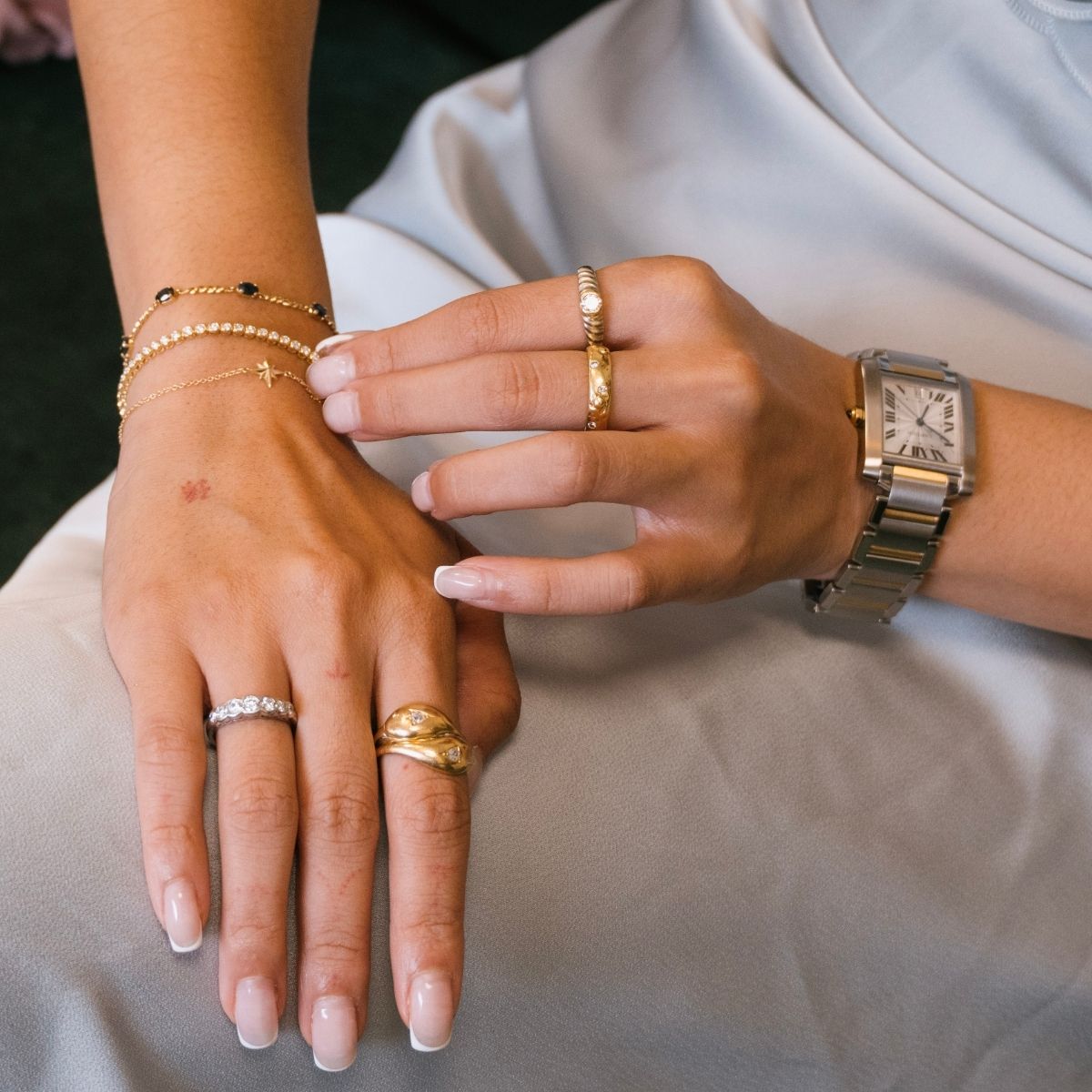 A close-up of a person's hands wearing multiple, vintage gold rings and preloved bracelets, with a two tone Cartier Tank watch on the wrist and a light blue satin dress in the background.