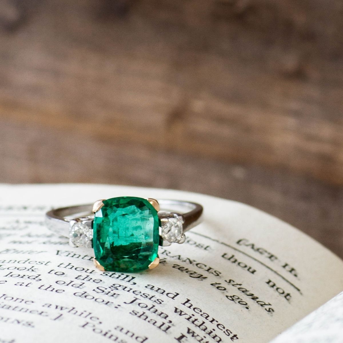 Close-up of a vintage emerald engagement ring with side accent diamonds, resting on an antique book.