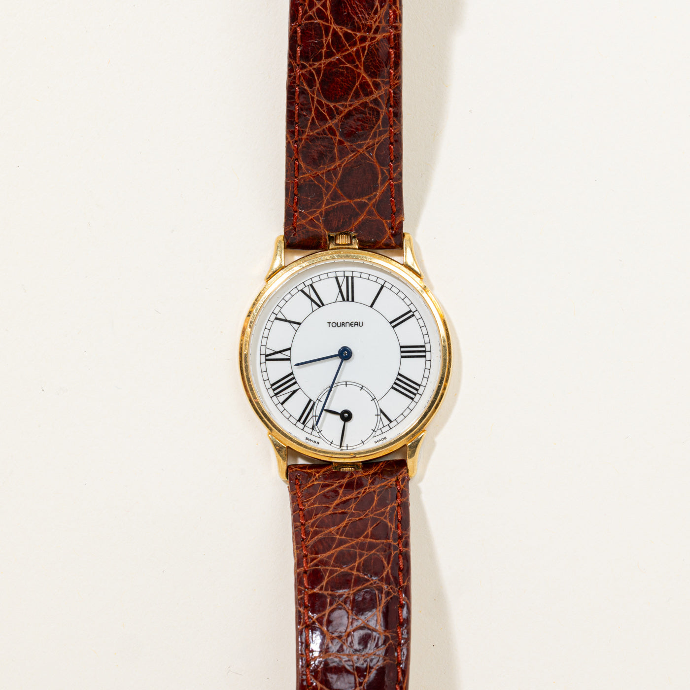 A vintage Tourneau wristwatch with a round gold case, white dial with black Roman numerals, and a brown textured leather strap, shown against a plain off-white background.