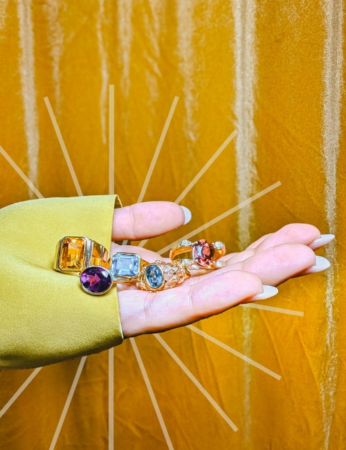 hand full of colourful gemstone rings in front of golden yellow backdrop with starburst graphic