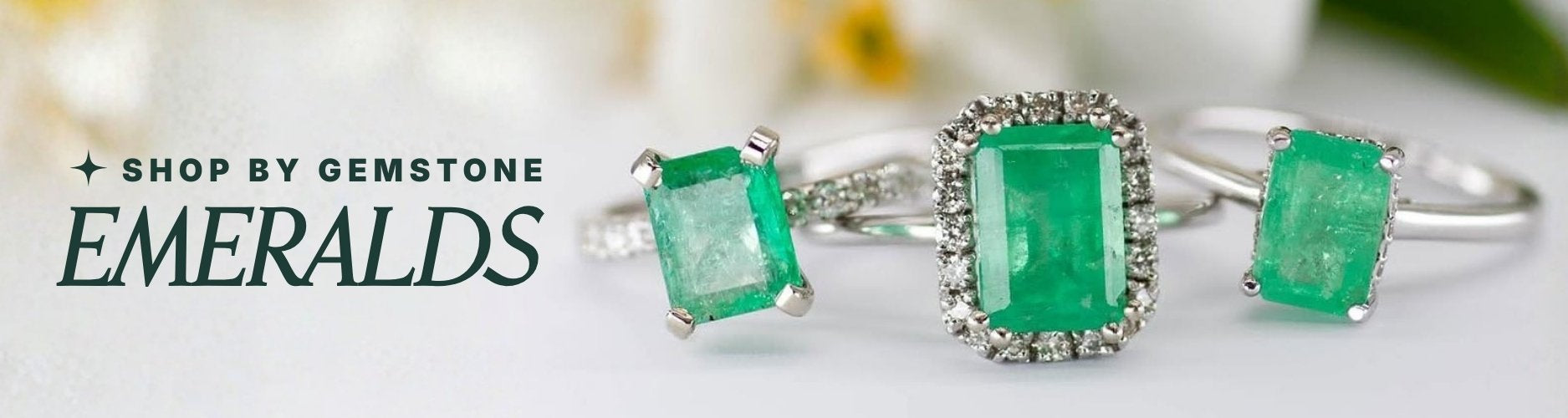 Three emerald rings in white gold settings on a white background with out of focus florals in background. Text overlay reads: 'Shop by Gemstone: Emeralds'