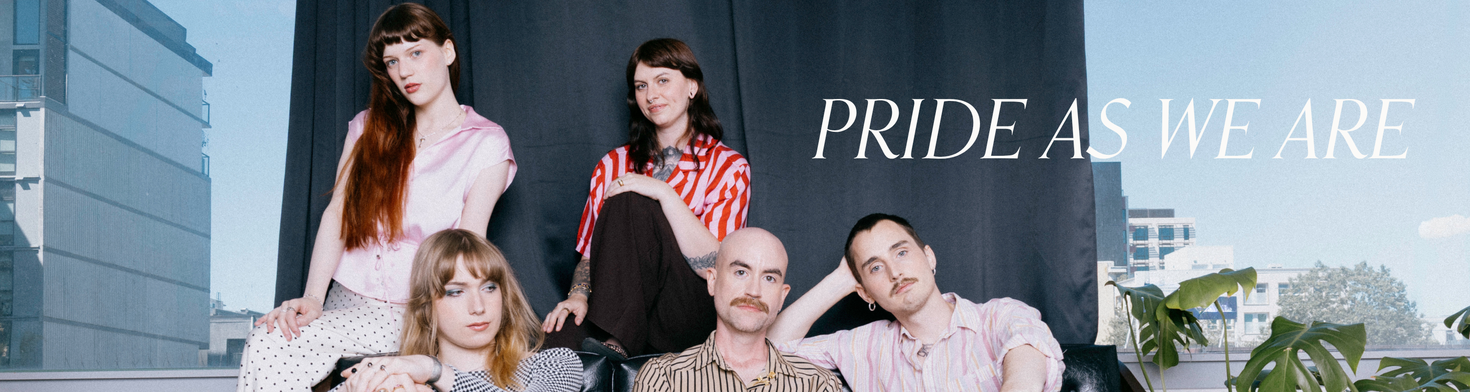 A group of five people posing together outdoors against a dark curtain backdrop with a blue sky and buildings in the background. White text on the right reads 'PRIDE as WE ARE'.