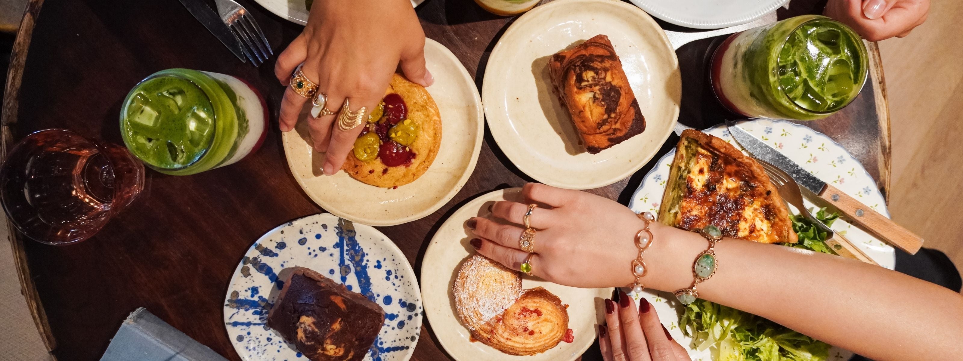 An overhead view of friends wearing unique, vintage gold and gemstone rings and bracelets while reaching for pastries and drinks at a cafe table.