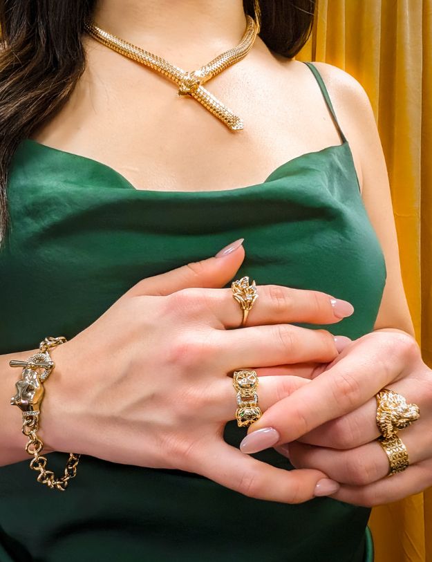 woman in green dress in front of golden yellow backdrop wearing gold snake necklace with ruby eyes and a selection of rings and bracelets featuring florals and animals from the 100 ways wildflower wildlife collection