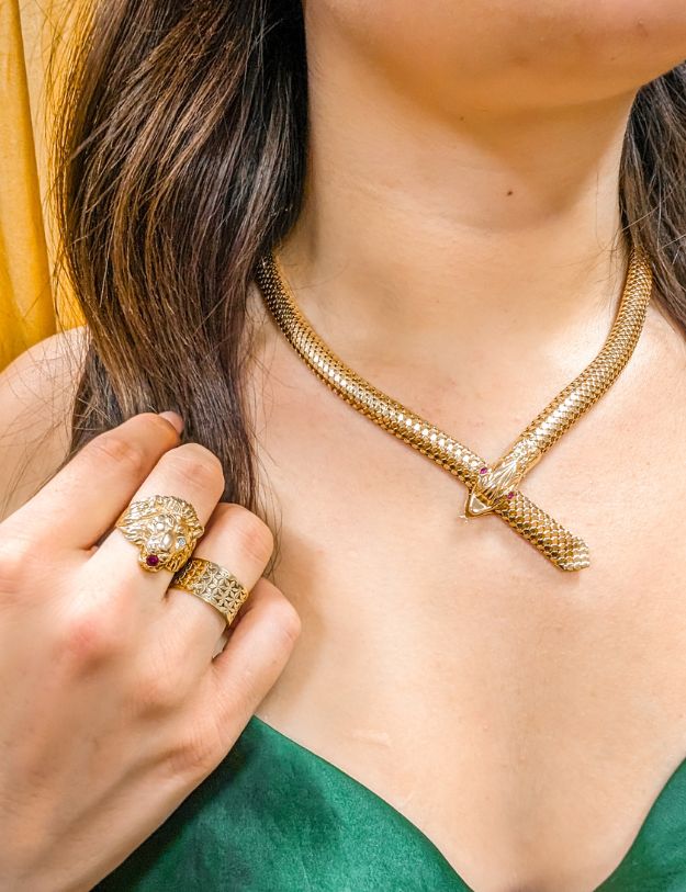 woman in green dress in front of golden yellow backdrop wearing gold snake necklace with ruby eyes and lion's face ring with diamond and ruby as well as a gold lattice ring all from the 100 ways wildflower wildlife collection